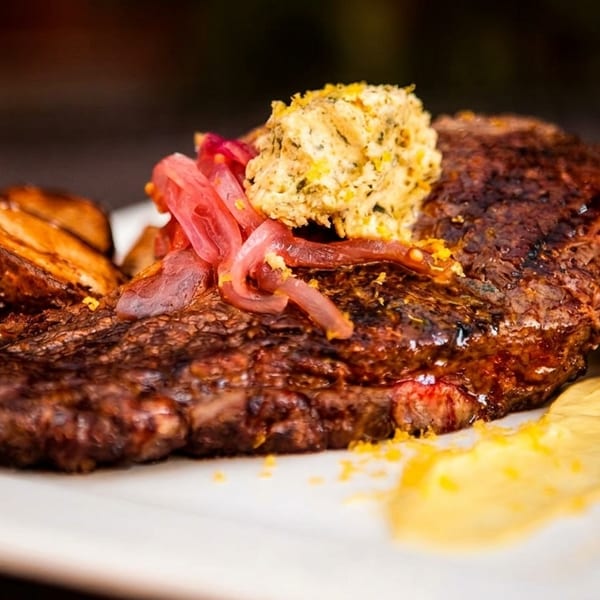 Grilled steak topped with herb butter, pickled onions, roasted potatoes, and a yellow sauce.