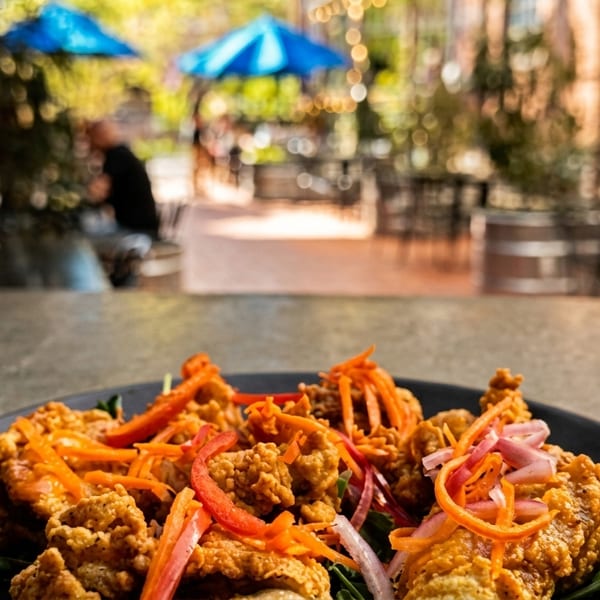 Crispy fried chicken with pickled vegetables on a black plate, with a blurred outdoor patio in the background.