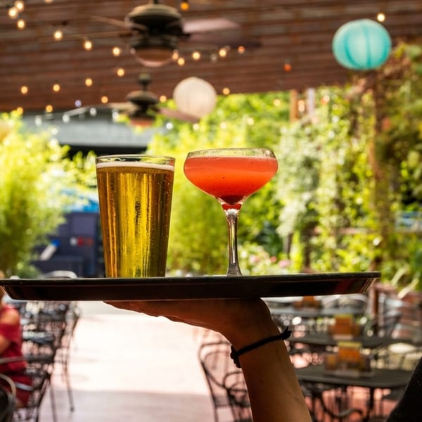 Server carrying a beer and a red cocktail on a tray through an outdoor patio.