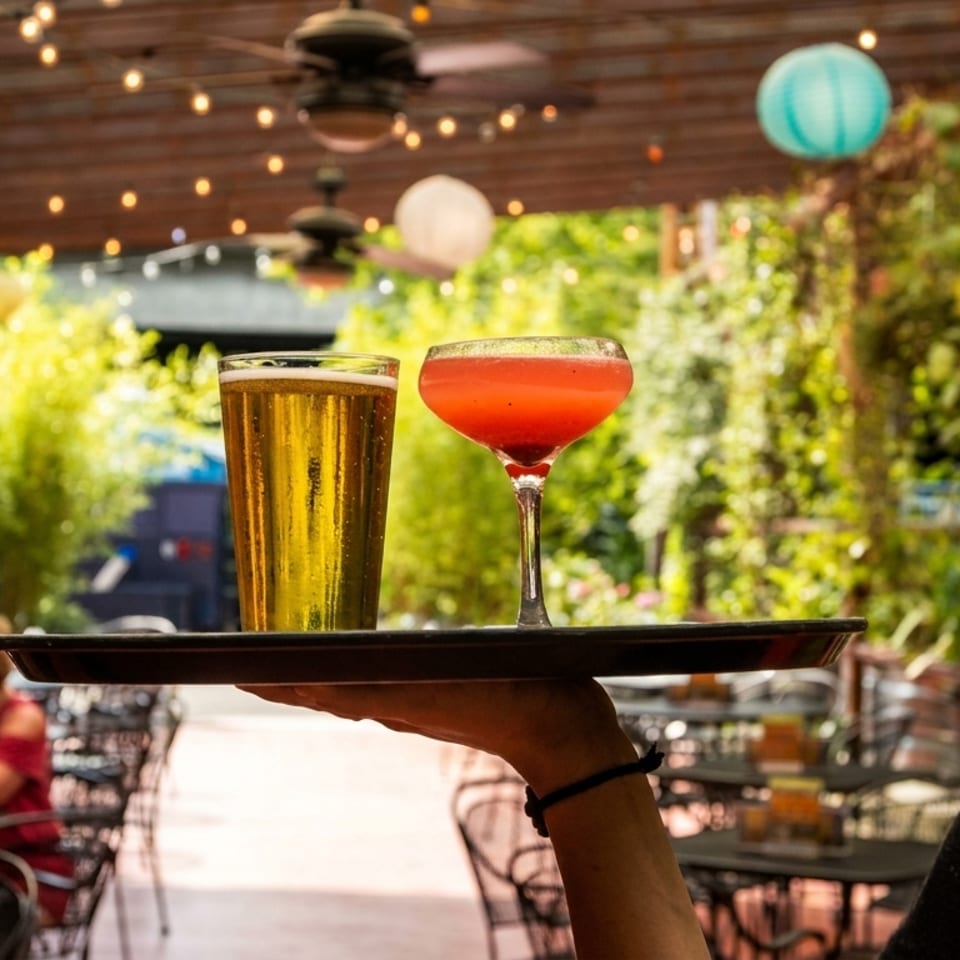 Server carrying a beer and a red cocktail on a tray through an outdoor patio with string lights.