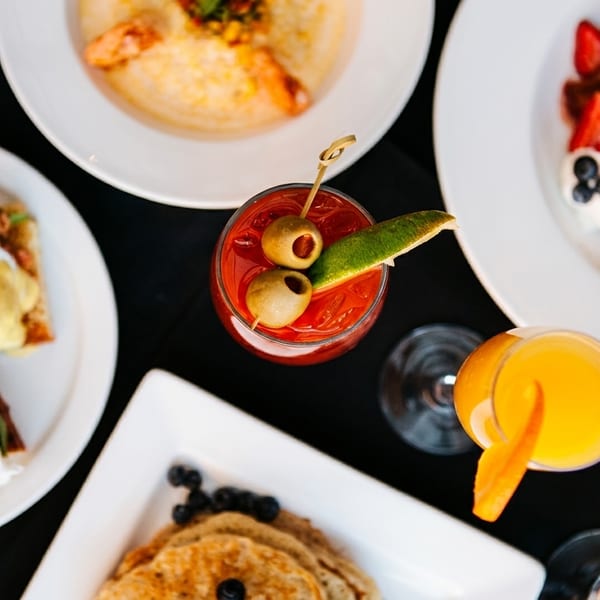 Overhead view of a brunch spread with Eggs Benedict, French toast, shrimp and grits, and cocktails.