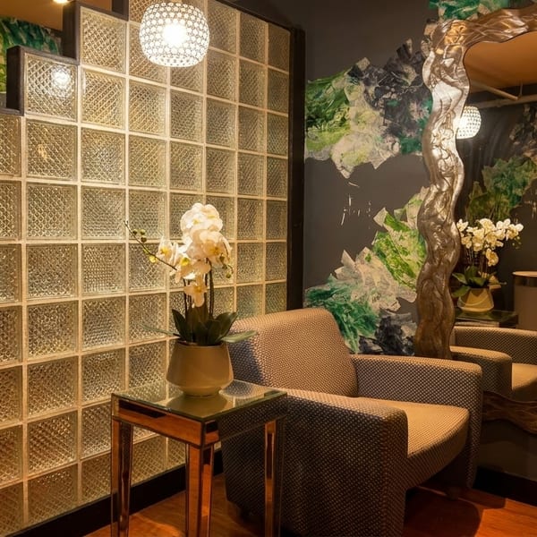 A cozy waiting area with a patterned armchair, glass block wall, and a large ornate mirror.